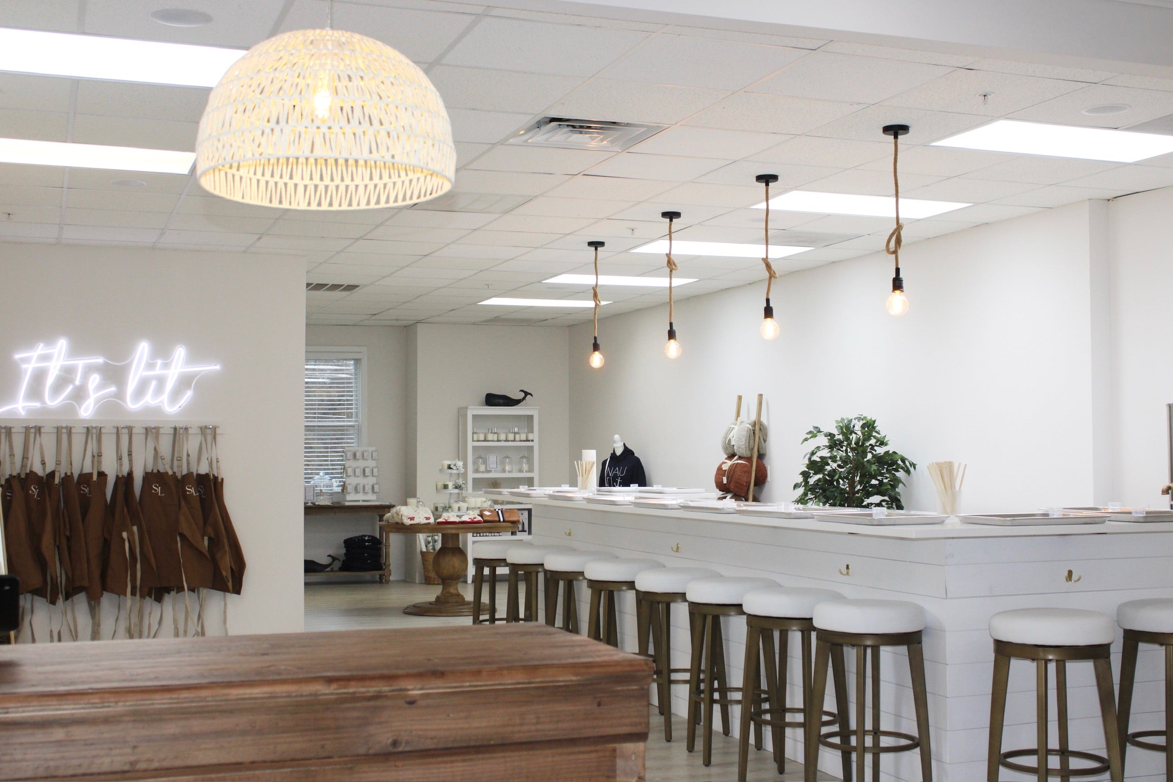 A modern and stylish interior of a candle boutique with minimalist design elements, featuring a white counter, chic bar stools, pendant lighting, and a neon sign on the wall.