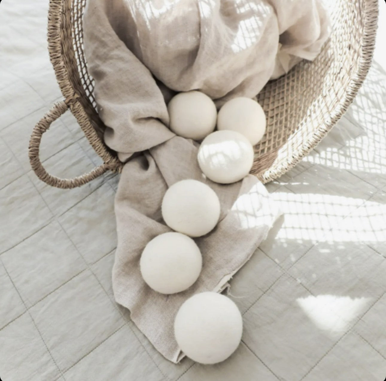 A collection of white decorative spheres elegantly spilling out of a woven basket onto a textured neutral fabric on the floor, bathed in soft, natural light.